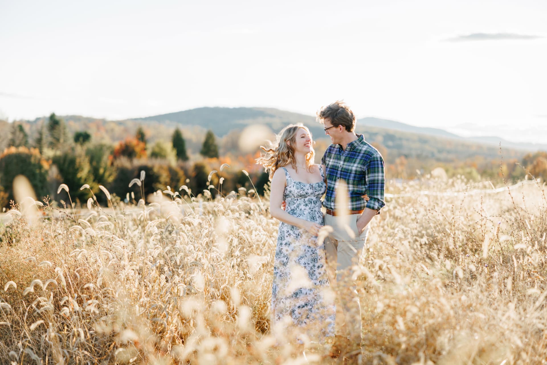 An engaged couple laughing together in a golden field with mountain views at golden hour